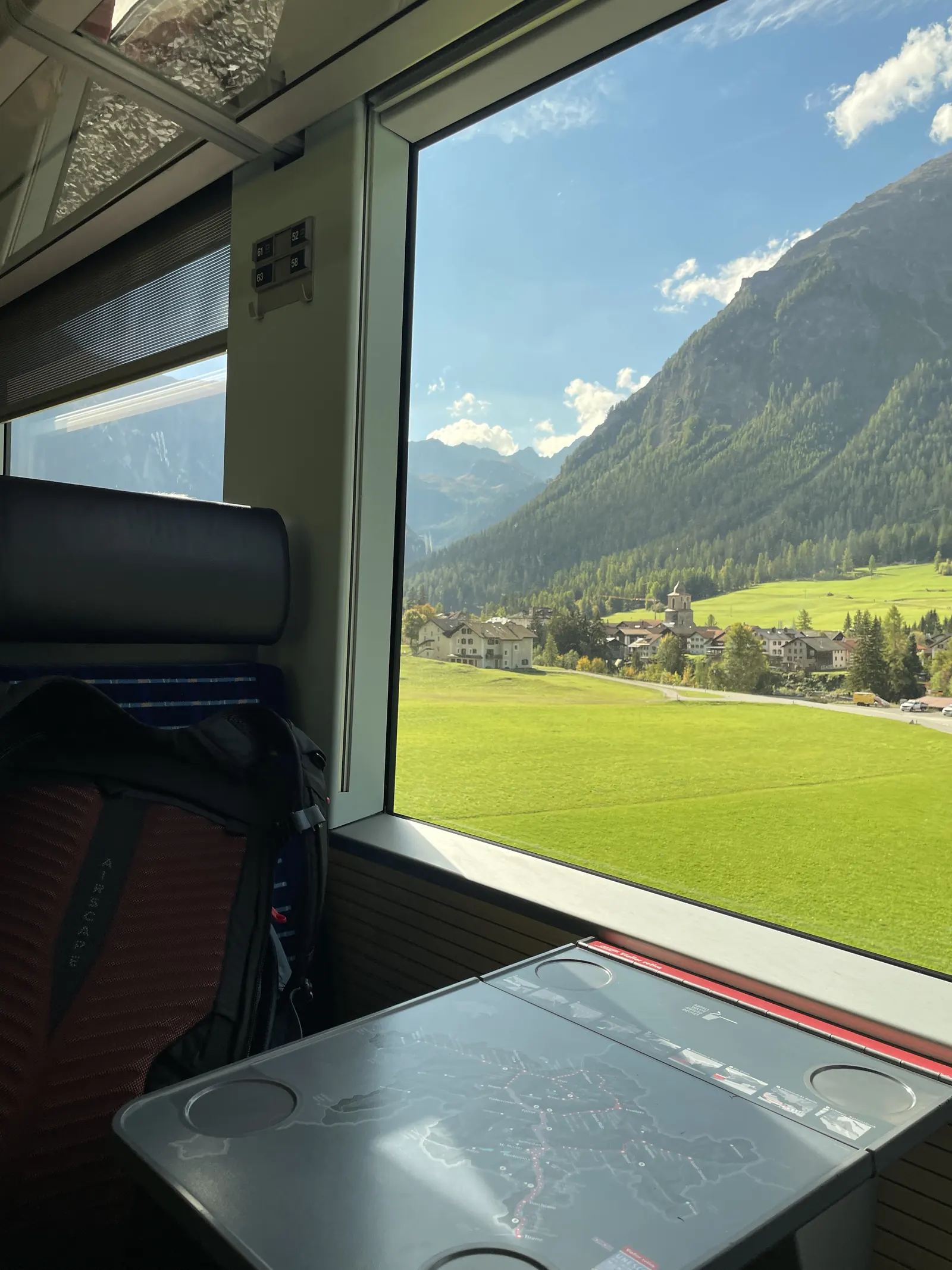Mountain scenery and a village seen through a train window, with a backpack beside the seat
