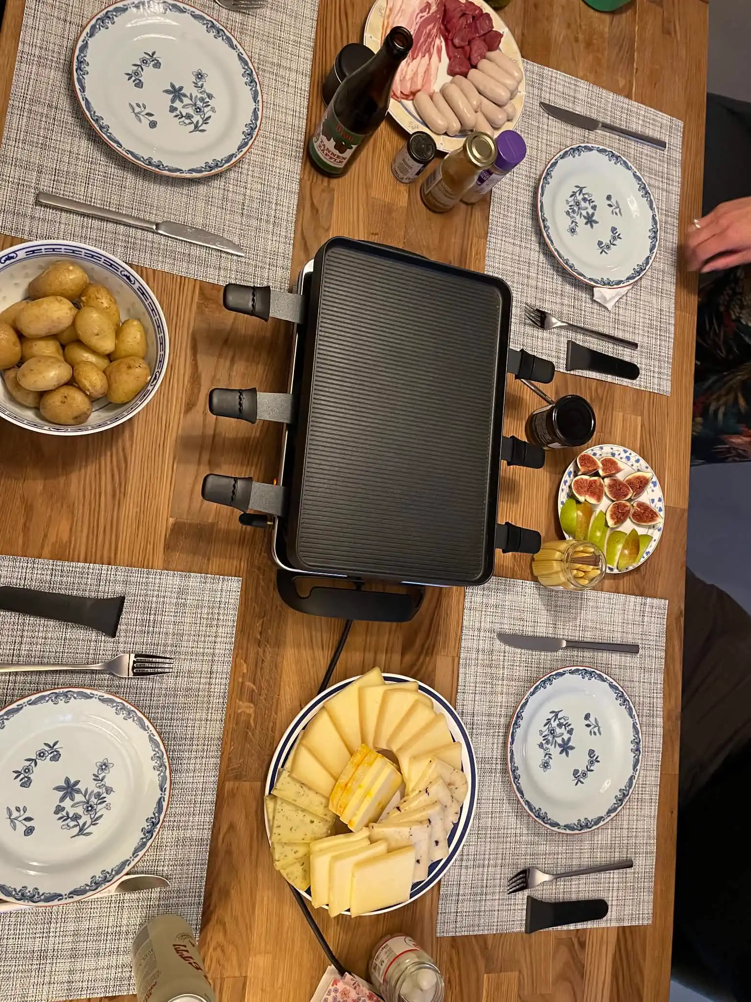 A home Swiss raclette meal with several cheeses, potatoes, meats, sausages, fruit, and a tabletop grill